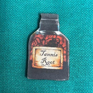 May include: A dark bottle-shaped image with a label that reads "Tannis Root." The bottle has a dark, shadowy appearance with red and orange accents. The label is rectangular with a tan background and elegant script.