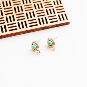 May include: A pair of gold-colored stud earrings with a turquoise-colored stone center. The earrings have a small loop at the bottom. A wooden, rectangular, openwork tray is in the background. The earrings are on a white surface.