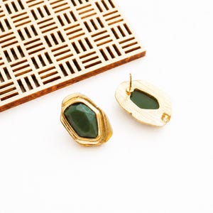 Peut inclure: Boucles d'oreilles à clous dorées avec une pierre verte foncée facettée. Les boucles d'oreilles ont une forme ovale irrégulière. Elles sont posées sur un fond blanc, avec un carré en bois à motifs géométriques en arrière-plan.