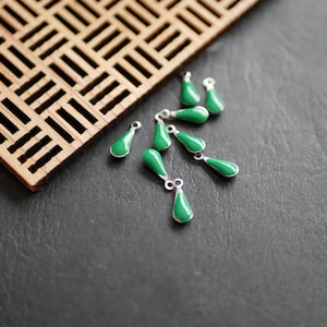 Peut inclure: Un groupe de huit breloques en forme de larme verte avec des boucles argentées. Les breloques sont disposées sur une surface noire.