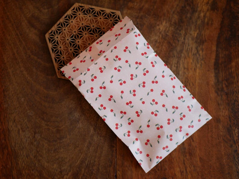 K&ouml;nnte beinhalten: Eine rosa-wei&szlig;e Stofftasche mit einem sich wiederholenden Muster aus roten Kirschen. Die Tasche ist gefaltet und liegt auf einer Holzoberfl&auml;che.