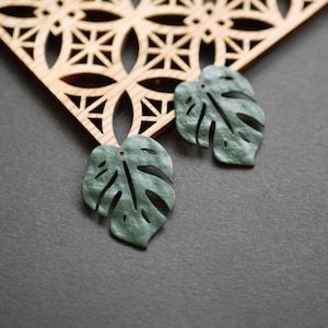 May include: Two green, leaf-shaped pendants with intricate cutouts, resembling Monstera leaves, rest on a dark gray surface. A wooden, geometric patterned element is in the background. The pendants have small holes for hanging.