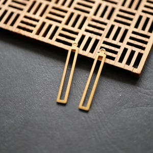 May include: Two gold-colored, rectangular earrings with a small loop at the top, resting on a dark gray surface. A wooden, geometric patterned piece is in the background. The earrings are a simple, modern design.