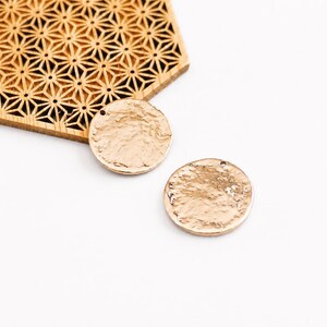 Peut inclure: Deux breloques circulaires en métal doré avec une surface texturée et martelée, dotées d'un petit trou sur le dessus pour la fixation, placées à côté d'une pièce en bois hexagonale avec un motif géométrique en étoile découpé au laser.