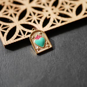 Peut inclure: Un pendentif doré avec un cœur turquoise et des flammes roses. Le pendentif a une forme arquée avec une bordure décorative. Il est posé sur une surface gris foncé, près d'une pièce décorative en bois.