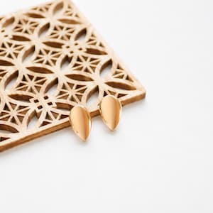 May include: A pair of polished rose gold teardrop-shaped stud earrings with a smooth, reflective surface. The earrings are positioned on a square wooden coaster featuring a laser-cut geometric pattern of intersecting circles and stars.