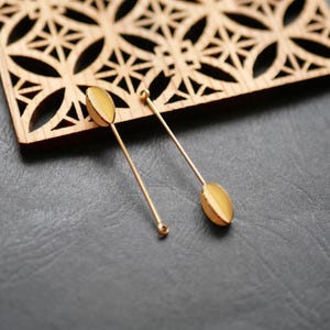 Pode incluir: Dois alfinetes de lapela dourados com um design oval, repousando sobre uma superfície cinza escuro. Uma peça decorativa de madeira com um padrão geométrico está no fundo. Os pinos têm uma haste longa e fina.
