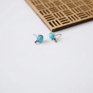 May include: A pair of stud earrings with a round, light blue stone set in a silver-colored metal. The earrings have a small loop detail below the stone. The background is white with a wooden decorative element.