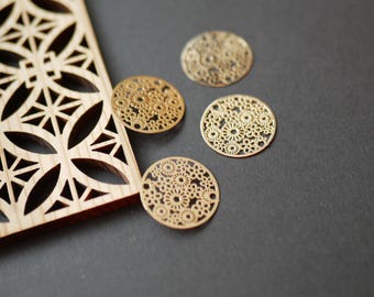 May include: Four gold-colored, circular metal charms with intricate floral cut-out designs. A wooden piece with a geometric pattern is on the left. The charms are on a dark gray surface.