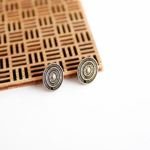 Può includere: Un paio di orecchini a lobo di forma ovale con un intricato motivo a cerchi concentrici in finitura argento antico. Ogni orecchino presenta un piccolo punto centrale in rilievo e una superficie testurizzata con sottili linee radiali.