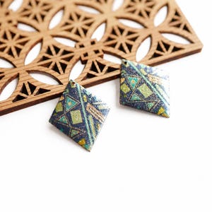 Peut inclure: Deux boucles d'oreilles en forme de losange avec une base bleu foncé et un motif géométrique composé de lignes et de formes vertes, vert clair et dorées. La surface présente une texture légèrement pailletée et un petit trou en haut.
