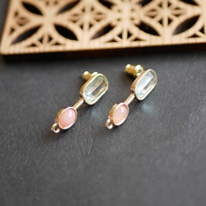 May include: A pair of gold-toned earrings with a light blue rectangular stone and a pink oval stone. The earrings are on a dark gray surface, with a wooden decorative piece in the background.