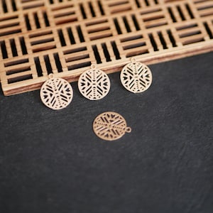 Op de afbeelding: Vier goudkleurige, cirkelvormige hangers met ingewikkelde geometrische uitsparingen. Eén hanger is geïsoleerd, terwijl de andere zich in de buurt van een houten, rechthoekige lade met een vergelijkbaar geometrisch patroon bevinden. De hangers hebben kleine lussen.