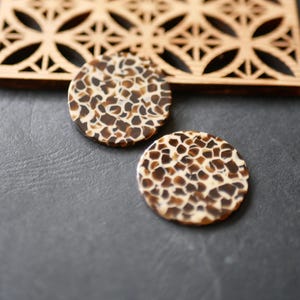 Peut inclure: Deux pendentifs ronds et plats avec un motif tacheté marron et crème. Un élément décoratif en bois avec un motif géométrique découpé est en arrière-plan. Les pendentifs sont sur une surface gris foncé.