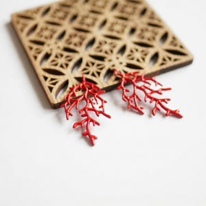 Peut inclure: Une paire de boucles d'oreilles rouges en forme de corail posées sur un dessous de verre en bois avec un motif géométrique. Les boucles d'oreilles sont d'un rouge vif et ont un design délicat et ramifié.