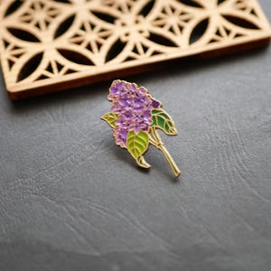 以下が含まれることがあります： 金色のアクセントが付いた、紫と緑のエナメルライラックの花のピン。 ピンは、緑の葉と金の茎を持つライラックの花の詳細なデザインが特徴です。 ピンは濃い灰色の背景に設定されています。