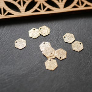Puede incluir: Pequeños dijes hexagonales dorados con una superficie texturizada y un solo agujero en la parte superior. Los dijes están esparcidos sobre una superficie gris oscuro, con un elemento decorativo de madera en el fondo.