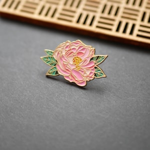Può includere: Una spilla a forma di fiore in smalto rosa e oro con foglie verdi. La spilla presenta un design dettagliato di una peonia. La spilla è posta su uno sfondo grigio scuro, con un elemento decorativo in legno sullo sfondo.