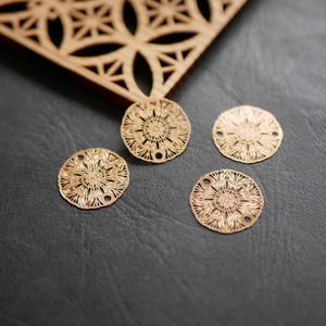 Peut inclure: Quatre breloques circulaires en métal doré, avec un motif floral ajouré, sont présentées sur une surface gris foncé. Les breloques ont deux petits trous pour l'enfilage. Une pièce décorative en bois découpée au laser est en arrière-plan.
