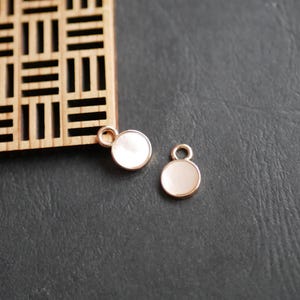 Op de afbeelding: Twee kleine, ronde bedels met een goudkleurige rand en een wit, parelmoerachtig midden. Elke bedel heeft een kleine lus aan de bovenkant om aan een ketting of ander sieradenonderdeel te bevestigen. De bedels liggen op een donkergrijze ondergrond.