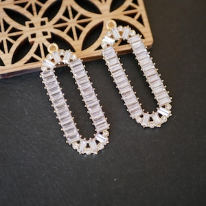Peut inclure: Deux composants de boucles d'oreilles ovales allongées de couleur dorée. Chaque boucle d'oreille est incrustée de cristaux clairs de coupe rectangulaire. Le haut et le bas de chaque boucle d'oreille présentent un motif de cristal décoratif.