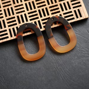 Peut inclure: Deux pendentifs de boucles d'oreilles ovales en acrylique, motif écaille de tortue. Les pendentifs présentent un dégradé de marron foncé à ambre. Chacun a un petit trou en haut pour l'attache des crochets. Ils sont présentés sur un treillis en bois.