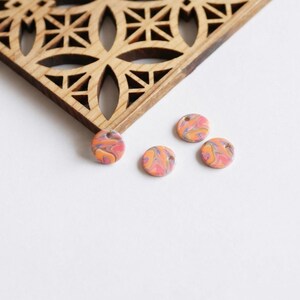 May include: Four small, round, marbled polymer clay beads in shades of pink, orange, and purple, each with a small hole. A wooden, geometric patterned square is in the background.
