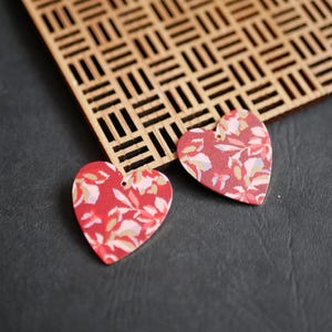 May include: Two heart-shaped earrings with a floral pattern in red, pink, and white. The earrings have small holes at the top. A wooden lattice is in the background, set against a dark gray surface.