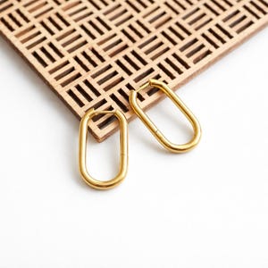 Peut inclure: Deux boucles d'oreilles créoles ovales dorées. Les boucles d'oreilles sont présentées sur une surface en bois clair avec un motif géométrique. Le fond est blanc, offrant un contraste net avec les bijoux.