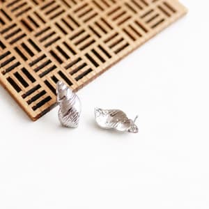 May include: A pair of silver-toned stud earrings shaped like small seashells. Each earring features a textured, spiralled surface with a metallic finish and a standard post backing, designed to resemble the natural form of a marine gastropod shell.