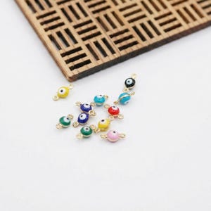 Puede incluir: Una colección de amuletos de ojo turco de esmalte colorido con herrajes dorados. Los amuletos presentan varios colores, incluyendo amarillo, verde, azul, rojo y rosa. Los amuletos son pequeños y redondos, con un diseño de ojo central.