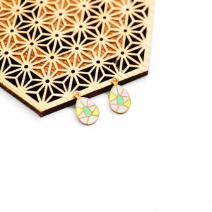Peut inclure: Deux pendentifs en forme de goutte d'eau avec des bords dorés et des motifs en émail coloré. Chaque pendentif présente une bordure blanche, avec des sections roses, jaunes et vertes. Les pendentifs sont présentés sur une surface en bois à motifs géométriques.