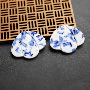 Può includere: Due ciondoli in ceramica bianca con un motivo floreale blu. I ciondoli sono a forma di petalo e hanno un foro nella parte superiore per l'infilatura. Sono esposti su una superficie scura con un reticolo di legno sullo sfondo.