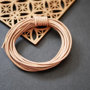 Könnte beinhalten: Ein aufgerolltes Bündel aus hellbraunem Lederband liegt auf einer dunkelgrauen Oberfläche, neben einem dekorativen Holzstück mit geometrischem Ausschnittmuster. Das Band ist eng gewickelt und zeigt seine Textur und Farbe.