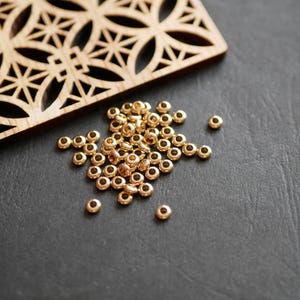 Può includere: Un insieme di piccole perline dorate sparse su una superficie scura. Un elemento decorativo in legno con un motivo geometrico è sullo sfondo. Queste perline sono adatte per progetti di creazione di gioielli e artigianato.