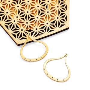 Può includere: Due componenti di orecchini a forma di goccia color oro con piccoli fori lungo la curva inferiore. Sullo sfondo c'è una forma geometrica in legno. Gli orecchini sono su sfondo bianco.