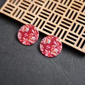 Può includere: Due orecchini rotondi rossi con un motivo floreale bianco, rosa e giallo. Sono posizionati su una superficie grigio scuro, vicino a un traliccio di legno. Bigiotteria artigianale.