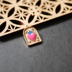 Peut inclure: Pendentif doré avec un cœur rose et des flammes bleues. Le pendentif est en forme d'arche avec des étoiles. Un élément décoratif en bois avec un motif géométrique est en arrière-plan.
