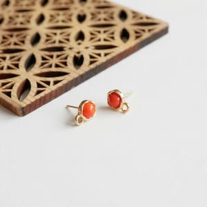 Puede incluir: Un par de pendientes de botón dorados con una pequeña gema redonda de color naranja engastada en un marco cuadrado. Los pendientes se exhiben sobre una superficie blanca, con un posavasos de madera decorativo en el fondo.