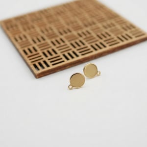 Puede incluir: Dos bases redondas doradas para pendientes con pequeños bucles, sobre un fondo blanco. En el fondo hay un posavasos cuadrado de madera con dibujos. Los pendientes son pequeños, probablemente para la fabricación de joyas.