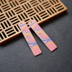 Puede incluir: Un par de pendientes rectangulares de color rosa, amarillo y azul con efecto marmoleado. Los pendientes están hechos de arcilla polimérica y tienen un gancho de metal dorado.