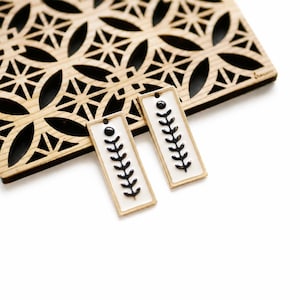May include: Two rectangular earrings with a white background and a black leaf design. The earrings have a gold border and a black dot at the top. A wooden decorative piece with a geometric pattern is in the background.