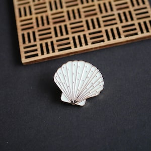 May include: A white and gold seashell enamel pin. The pin has a detailed design with radiating lines and small dot accents. It is set against a dark background with a wooden decorative element in the upper left corner.