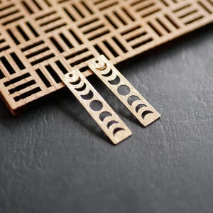 May include: A pair of gold-toned earrings with a moon phase design. The earrings are made of wood and have a rectangular shape.
