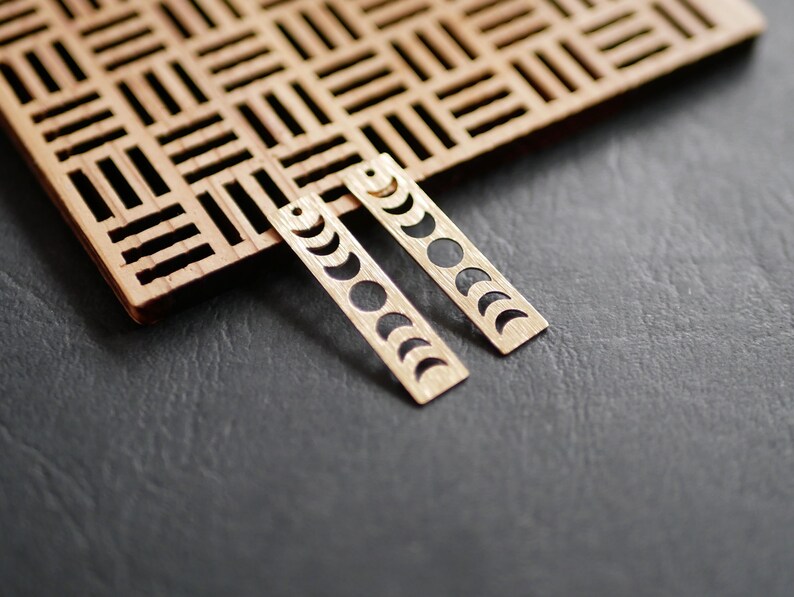 K&ouml;nnte beinhalten: Ein Paar goldfarbene Ohrringe mit einem Mondphasen-Design. Die Ohrringe sind aus Holz und haben eine rechteckige Form.