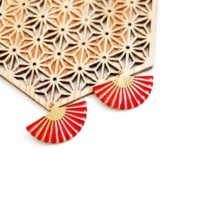 Könnte beinhalten: Ein Paar Ohrringe aus goldfarbenem Metall in Form von Miniatur-Faltfächern mit leuchtend roter Emaille-Verzierung, platziert vor einer hellen Holzplatte mit einem lasergeschnittenen geometrischen Sternmuster und sechseckiger Symmetrie.