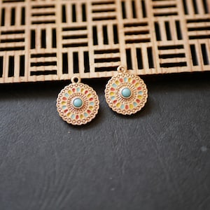 Peut inclure: Deux pendentifs ronds dorés avec un centre turquoise et des détails en émail coloré. Les pendentifs ont un design décoratif en forme de fleur. Ils sont présentés sur une surface sombre avec un treillis en bois en arrière-plan.