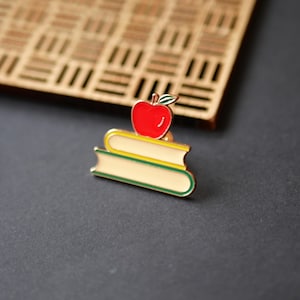 Op de afbeelding: Een kleurrijke emaille pin met een rode appel bovenop twee gestapelde boeken. De boeken hebben crèmekleurige pagina's met groene en gele accenten. De pin is waarschijnlijk een accessoire met een thema voor leraren of studenten.