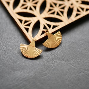 May include: Two gold-colored fan-shaped earrings with a textured, ridged design. The earrings are displayed on a gray surface, with a wooden decorative element in the background. The earrings have a small loop at the top for attachment.