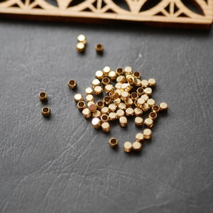 May include: A scattering of small, gold-coloured metal beads on a dark grey surface. The beads are cylindrical with a hole through the centre. A wooden decorative element is visible in the background.